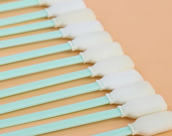 Puede incluir: Primer plano de una fila de 15 hisopos con punta de espuma blanca y mango azul. Los hisopos están dispuestos en diagonal sobre una superficie de color naranja claro.