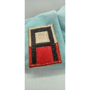 May include: A rectangular embroidered patch featuring a geometric design with a central beige square positioned above a red square, both framed by thick black vertical borders and a tan stitched edge, set against a light blue fabric background.