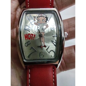 Puede incluir: Un reloj de pulsera plateado con una correa de cuero roja. La esfera del reloj presenta una imagen de dibujos animados de una enfermera con la palabra "HOT!" impresa en la esfera.
