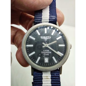 Könnte beinhalten: Eine Dakota-Uhr mit schwarzem Zifferblatt, weißen Stundenmarkierungen und silbernen Zeigern. Die Uhr hat ein Titangehäuse und eine Datumsanzeige. Das Uhrenarmband ist blau und weiß gestreift. Die Uhr wird von einer Hand gehalten.