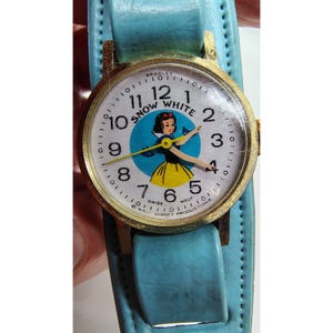 以下が含まれることがあります： ヴィンテージのスノーホワイトの腕時計。金色のケースと水色の革バンド。文字盤には青い円の中にスノーホワイトのキャラクターが描かれ、黒い数字と針があります。この時計はブラッドリー スイス製です。