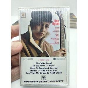 May include: A vintage cassette tape featuring Bob Dylan. The cassette tape is white with red and black text. The text reads "Bob Dylan" and "Featuring She's No Good In My Time Of Dyin' Man Of Constant Sorrow House Of The Risin' Sun See That My Grave Is Kept Clean". The cassette tape is labeled "Columbia Stereo Cassette".