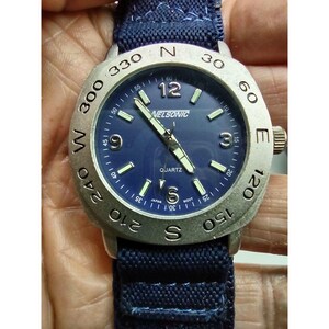 May include: A silver watch with a blue face and a black canvas band. The watch face has white markings and the text "NELSONIC" and "QUARTZ".
