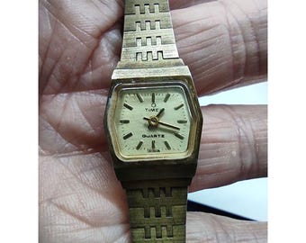 May include: A gold-toned wristwatch with a square face and a gold metal band. The watch face has black hour and minute hands, a black second hand, and a white background with the words "Q Time Quartz Taiwan" printed on it.