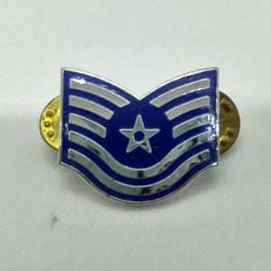 Peut inclure: Une épingle en métal bleu et argenté avec une étoile au centre. L'épingle a un fond bleu avec des rayures argentées et un bord argenté. L'épingle est fixée avec deux attaches dorées.