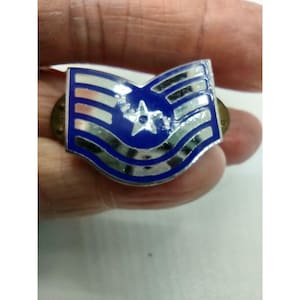 Peut inclure: Une épinglette d'insigne militaire bleue et argentée avec une étoile au centre. L'épinglette a un fond bleu avec des rayures argentées et une bordure argentée. L'épinglette est tenue dans une main.