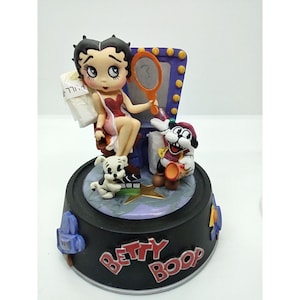 以下が含まれることがあります： ベティ・ブープのフィギュア。ベティ・ブープは赤いドレスを着て、鏡を持ち、化粧台の鏡の前に座っています。犬と子犬もいます。ベースは黒で、「Betty Boop」の文字が赤色で書かれています。