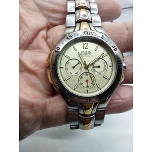 May include: A silver and gold two-tone Guess Waterpro watch with a white face and three subdials. The watch has a date window, day of the week display, and a 24-hour display. The watch is water resistant to 165 feet.