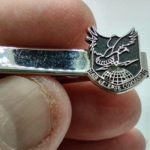 Puede incluir: Un pasador de corbata plateado con un emblema negro y plateado. El emblema representa un águila sobre un globo terráqueo, con el texto "1st AIR DEFENSE COMMAND". El pasador se sostiene entre los dedos, mostrando su superficie pulida.