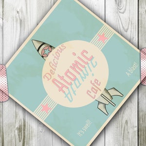 Puede incluir: Un póster de estilo retro con fondo azul y un círculo blanco en el centro. El círculo tiene el texto "Delicious Atomic Atomic Cafe" en letras rosa y blancas. Una nave espacial con una estrella rosa en el lateral está volando a través del círculo. El póster tiene el texto "It's swell!" y "A blast!" en las esquinas inferiores.