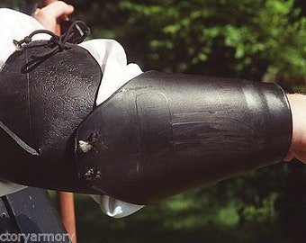 May include: Black leather arm armor, likely for historical reenactment or costume use. The armor is worn over a white shirt, with a textured surface and visible stitching. The design includes a rounded elbow piece and a forearm guard.
