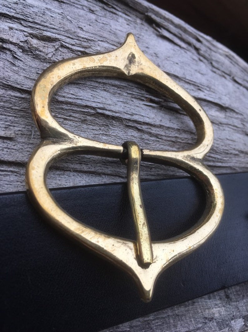 Pode incluir: Uma fivela de cinto de metal dourado com um design &uacute;nico e ornamentado. A fivela tem duas al&ccedil;as e uma parte superior pontiaguda.