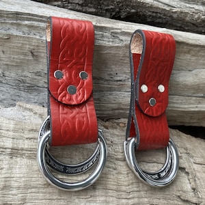 May include: Two red leather belt loops with silver metal rings. The leather has a textured pattern and is secured with silver rivets. The rings have a decorative etched design.