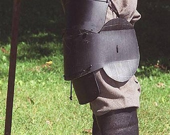 May include: A close-up of a person's leg wearing black leather knee pads and boots. The knee pads are secured with straps and buckles. The boots are tall and have a pointed toe.