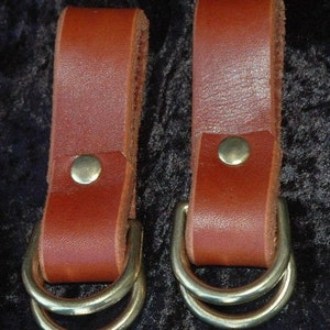 May include: Two brown leather straps with brass double D-rings. The straps are about 2 inches wide and have a single brass rivet on each.