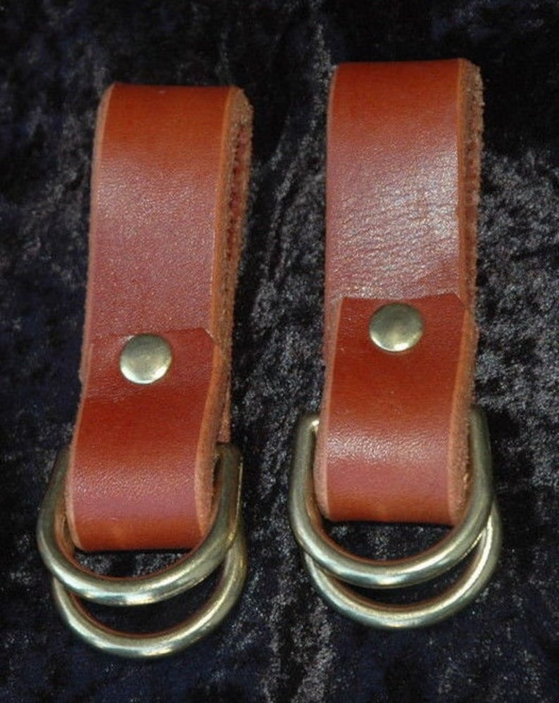 May include: Two brown leather straps with brass double D-rings. The straps are about 2 inches wide and have a single brass rivet on each.
