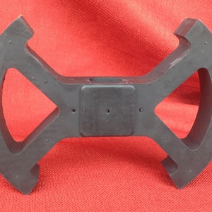 May include: Black plastic part with a central hole and four arms with cutouts. The arms are curved and have a series of small holes.