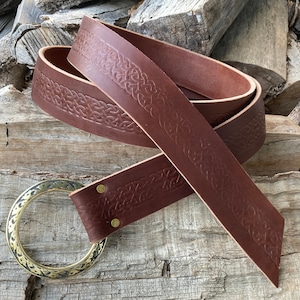 May include: A brown leather belt with a Celtic knot design embossed along the edges. The belt has a brass-colored circular buckle. The leather is a rich, dark brown color, and the belt is displayed on a wooden surface.