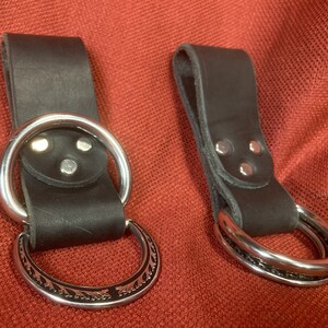 May include: Two black leather belt loops with silver metal rings. The loops have three silver rivets on the back. One ring is plain and the other has a decorative design.