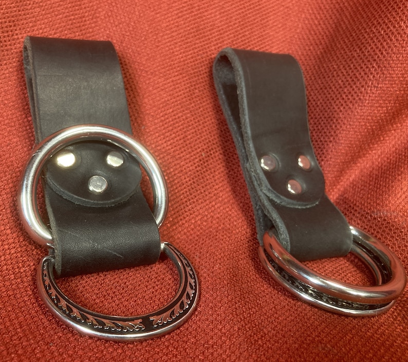 May include: Two black leather belt loops with silver metal rings. The loops have three silver rivets on the back. One ring is plain and the other has a decorative design.