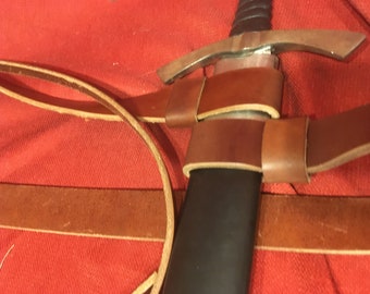 Peut inclure: Un fourreau d'épée en cuir marron avec une poignée en métal et une lame noire. Le fourreau est sécurisé par deux sangles.