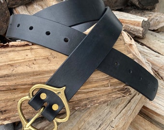 May include: A black leather belt with a decorative gold-colored buckle. The belt has several holes for adjustment and is displayed on a wooden surface. The buckle has an ornate, heart-shaped design.