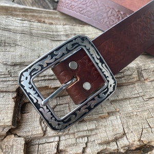 May include: A brown leather belt with an intricate embossed design and a silver buckle with a decorative border.