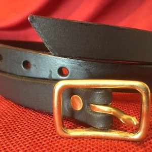 May include: A black leather belt with a rectangular gold-colored buckle. The belt has several evenly spaced holes and a pointed end. The buckle is a simple, classic design. The belt is shown against a red background.