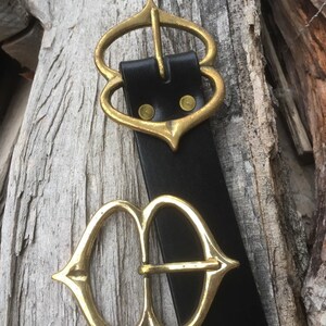 Pode incluir: Um cinto de couro preto com duas fivelas de metal dourado ornamentadas. As fivelas s&atilde;o ambas em forma de cora&ccedil;&atilde;o com uma ponta pontiaguda na parte inferior.