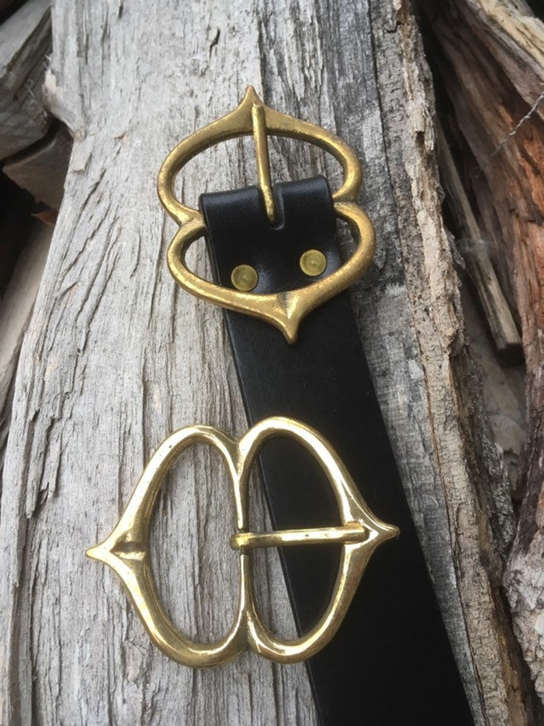 Pode incluir: Um cinto de couro preto com duas fivelas de metal dourado ornamentadas. As fivelas s&atilde;o ambas em forma de cora&ccedil;&atilde;o com uma ponta pontiaguda na parte inferior.