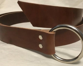 Peut inclure: Une ceinture en cuir marron avec une grande boucle en argent en forme d'anneau. La ceinture est pliée en deux et la boucle est sur le côté droit de l'image.