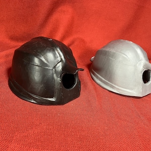 Könnte beinhalten: Zwei Helme, einer schwarz und einer silber, mit einem Loch oben und einem kleinen Flügel an der Seite.