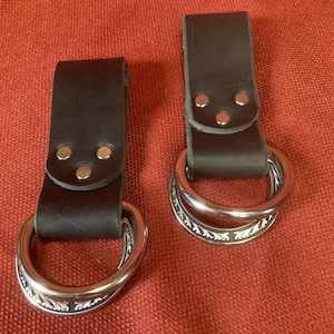 May include: Two brown leather belt loops with silver metal rings. The rings have a decorative design.