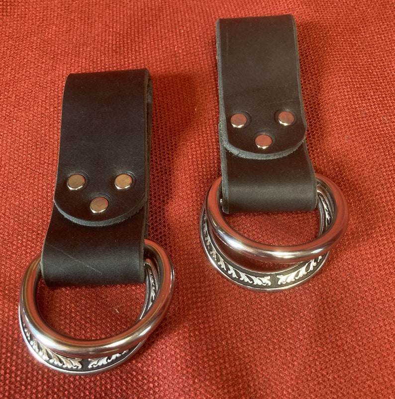 May include: Two brown leather belt loops with silver metal rings. The rings have a decorative design.