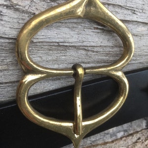 Pode incluir: Uma fivela de cinto de metal dourado com um design &uacute;nico e ornamentado. A fivela tem uma parte superior pontiaguda e duas al&ccedil;as arredondadas.