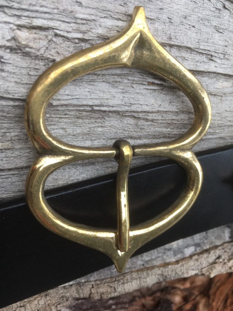 Pode incluir: Uma fivela de cinto de metal dourado com um design &uacute;nico e ornamentado. A fivela tem uma parte superior pontiaguda e duas al&ccedil;as arredondadas.