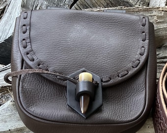 May include: A brown leather pouch with a flap closure and a decorative antler toggle. The pouch has a stitched design around the edges.
