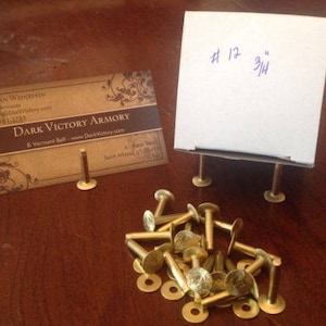 May include: A close-up of a pile of brass-colored metal rivets, with a business card for Dark Victory Armory and a white box labeled "#12 3/4" in the background.