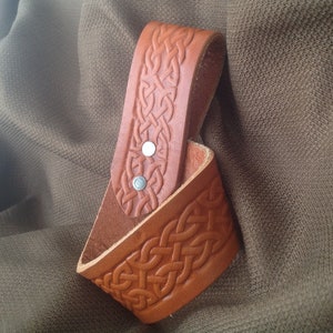 May include: A brown leather belt loop with a Celtic knot design. The loop has two silver rivets.