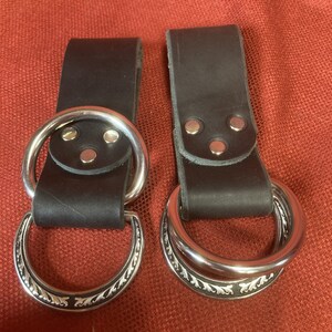 May include: Two brown leather belt loops with silver metal rings. The rings have a decorative design.