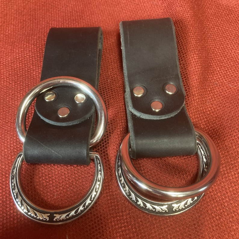 May include: Two brown leather belt loops with silver metal rings. The rings have a decorative design.