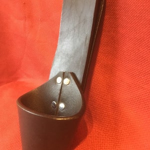 May include: A dark brown leather cup holder with a long strap, attached with silver rivets. The cup holder is designed to be worn on a belt. The background is a solid red color.