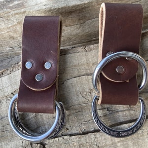 May include: Two brown leather belt loops with metal rings. One ring is plain and the other has a decorative design.