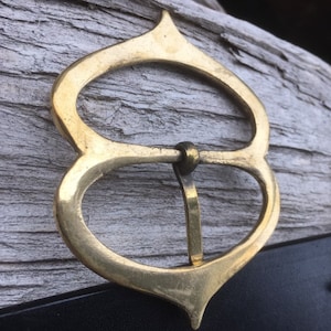 Pode incluir: Uma fivela de cinto de metal dourado com um design &uacute;nico e ornamentado. A fivela tem a forma de uma l&aacute;grima com um oval arredondado no centro. A fivela est&aacute; presa a um cinto de couro preto.