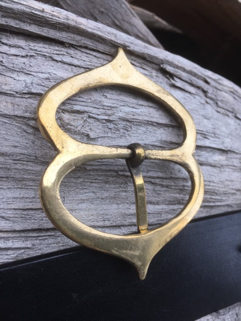 Pode incluir: Uma fivela de cinto de metal dourado com um design &uacute;nico e ornamentado. A fivela tem a forma de uma l&aacute;grima com um oval arredondado no centro. A fivela est&aacute; presa a um cinto de couro preto.