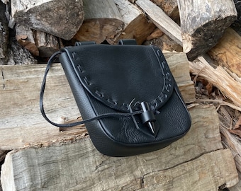 May include: A black leather pouch with a decorative flap and a pointed metal closure. The pouch has a strap for carrying it over the shoulder.