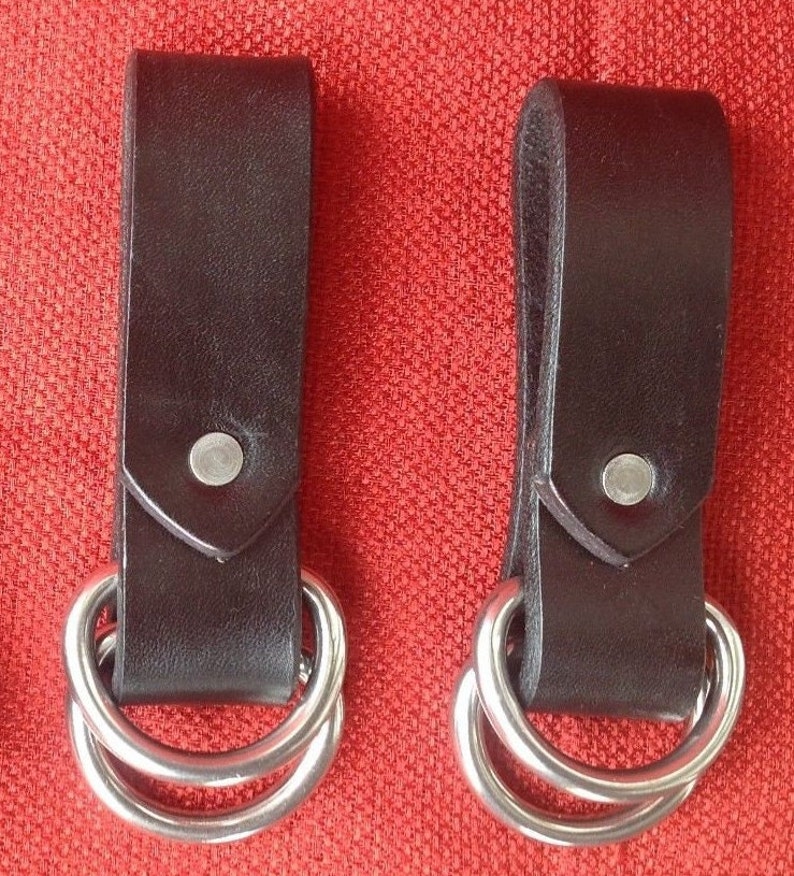 May include: Two black leather belt loops with silver double rings. The loops are on a red background.