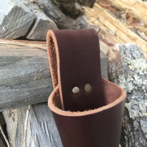 May include: A brown leather cup holder with two brass rivets. The holder is designed to hang from a belt or hook.