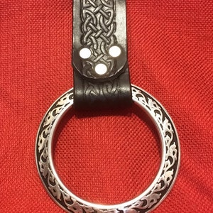 Könnte beinhalten: Eine schwarze Ledergürtelschlaufe mit einem keltischen Knotendesign und drei silbernen Nieten. Die Schlaufe ist an einem silbernen Ring mit einem dekorativen Blumenmuster befestigt.