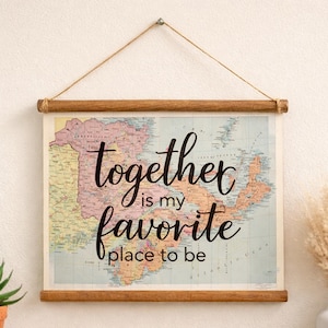 May include: A hanging wall art piece featuring a vintage-style map of the Canadian Maritimes with the black cursive text "together is my favorite place to be" printed over it. The print is held by two wooden bars and hangs from a twine string.